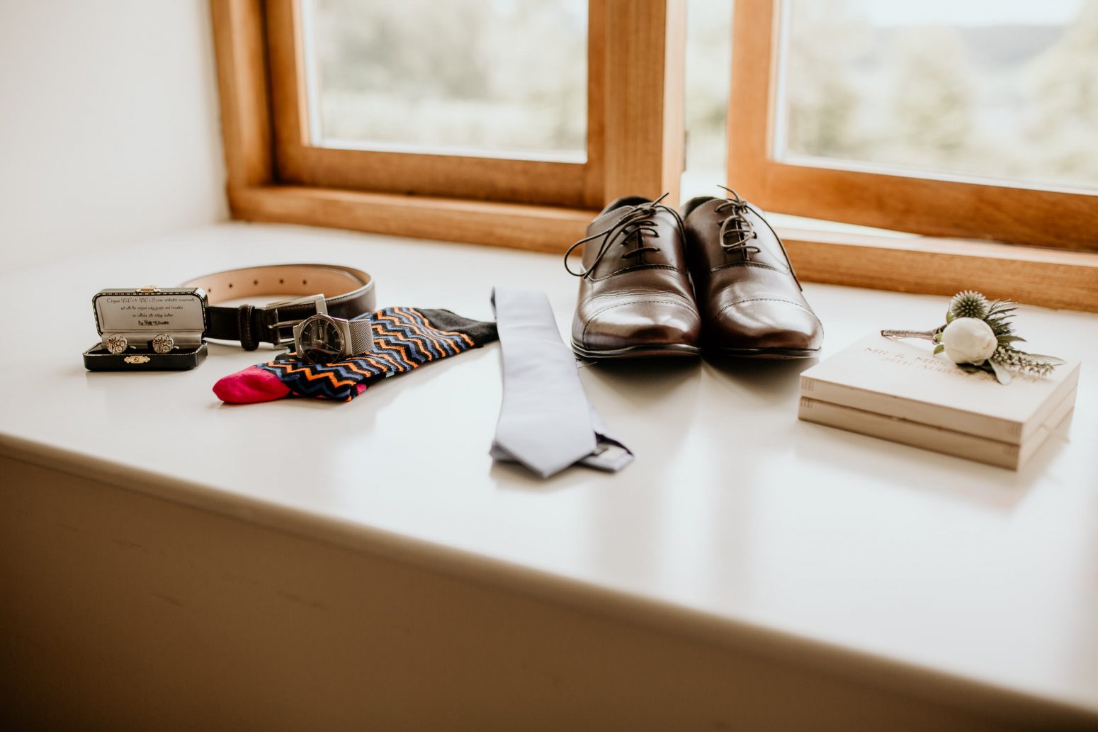 bright photo showing grooms wedding accessories on the morning of the wedding laid out on a bright windowsill including groom's shoes, belt, socks, cufflinks, watch and tie