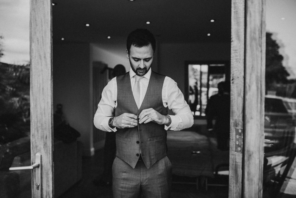 black and white photo of groom doing up grey waistcoat whilst walking through doorway