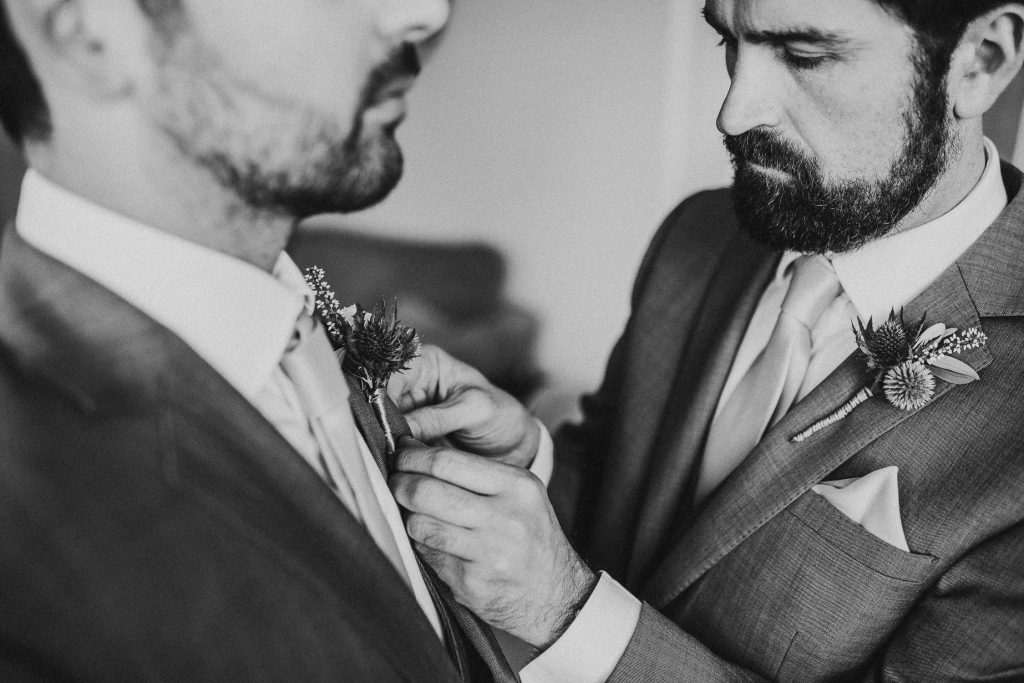 black and white close up of groomsman and groom in matching grey suits with light blue ties and green and white buttonholes