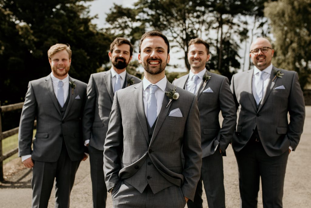 smiling groom and groomsmen in matching luxurious grey suits, waistcoats, light blue tie and green and white buttonholes stand outside in the sunshine