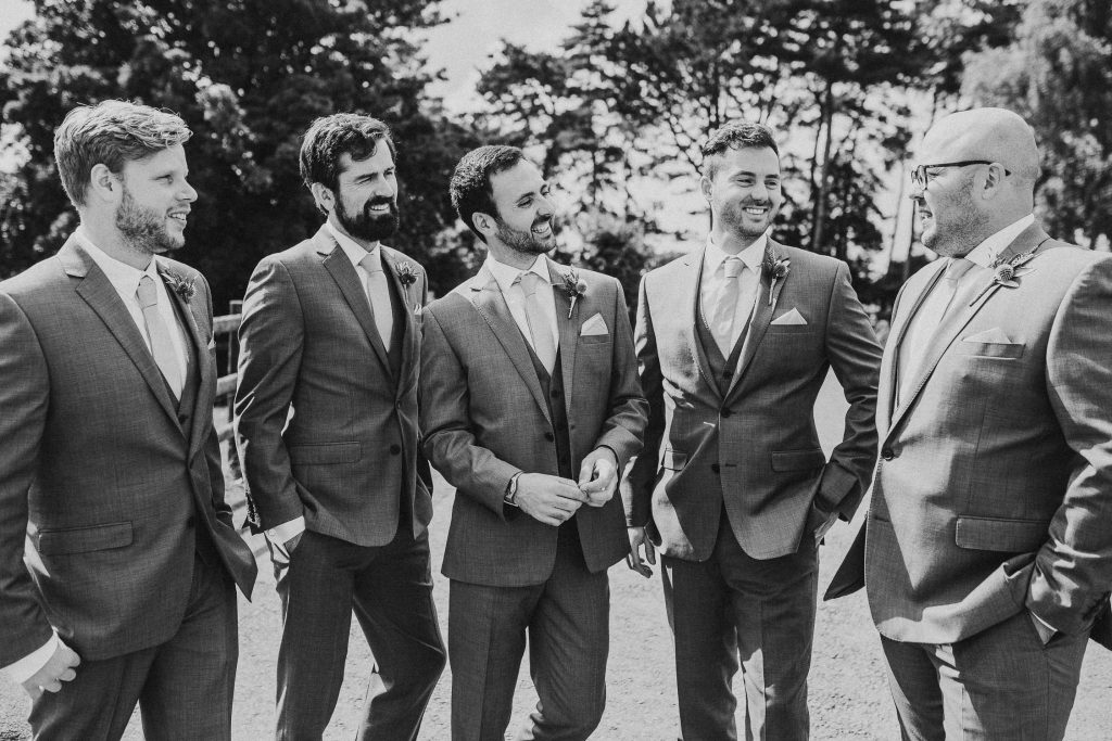 black and white photo of smiling groom and groomsmen in matching luxurious grey suits laugh together outside in the sunshine