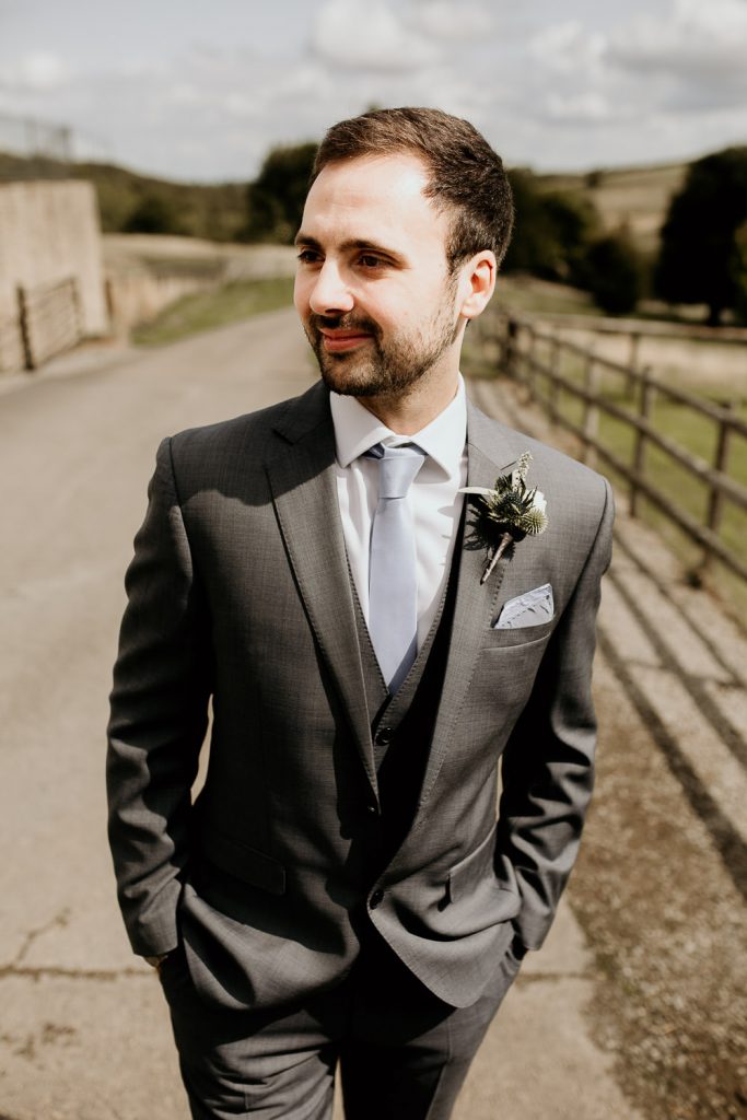 groom in grey suit, light blue tie and green and white buttonhole stands outside in the sunshine