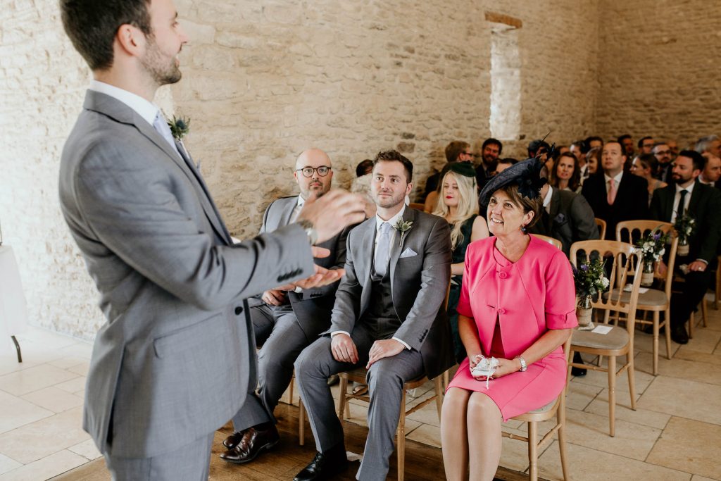 groom in grey suit stands at the alter smiling with groomsmen and wedding guests watching in luxurious barn conversion wedding