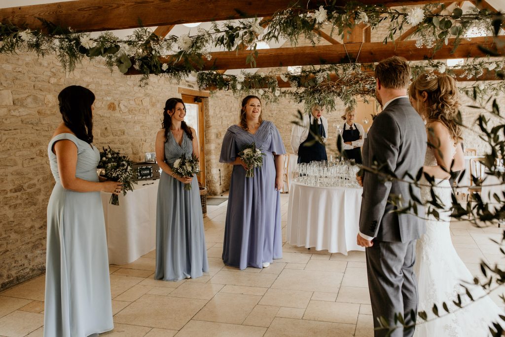 bride and father of the bride talk with bridesmaids in luxury barn conversion wedding venue with beams covered in fairy lights, white flowers and green foliage