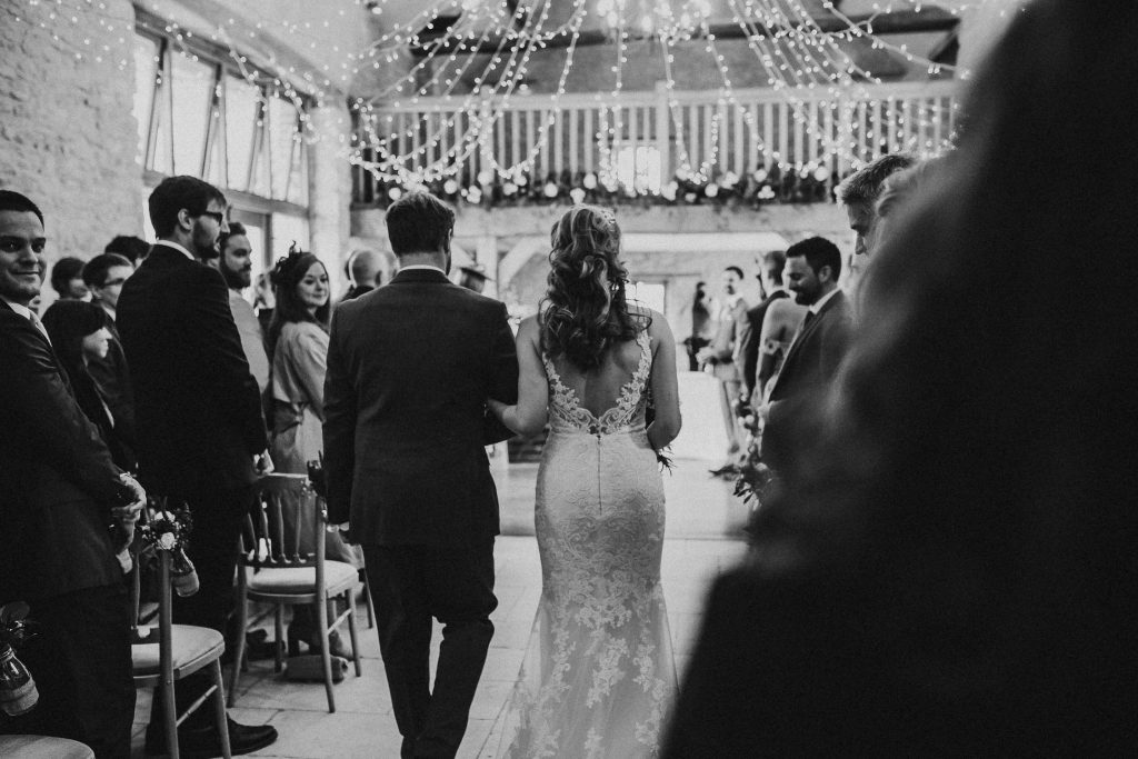black and white photo of bride being walked down the aisle past wedding guests in luxurious barn conversion wedding venue with fairy lights overhead