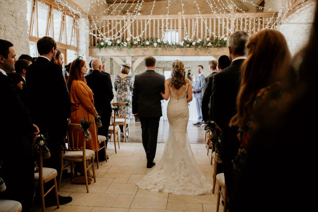 bride being walked down the aisle past wedding guests in luxurious barn conversion wedding venue with fairy lights overhead