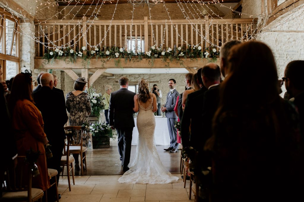 bride being walked down the aisle past wedding guests in luxurious barn conversion wedding venue with fairy lights overhead