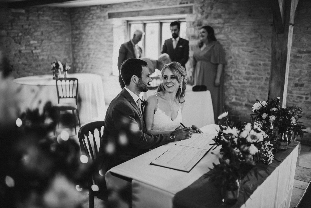 black and white bride smiles at groom as they sign the marriage register in luxurious beamed barn conversion wedding venue