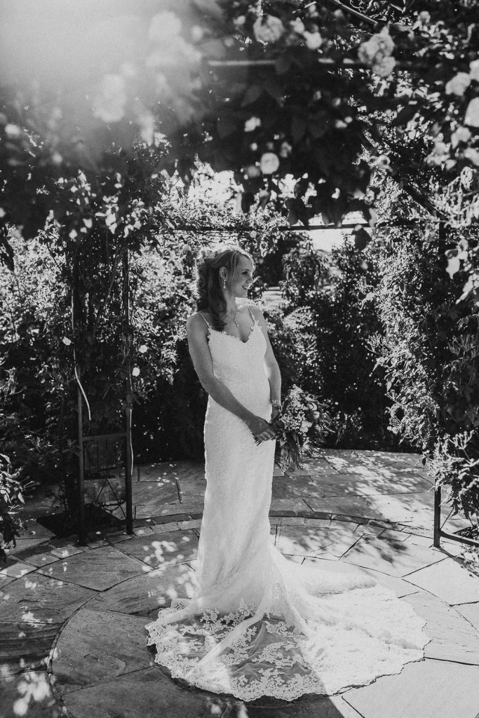 black and white photo of bride in luxurious white lace wedding dress with bouquet standing surrounded by trees and rich foliage