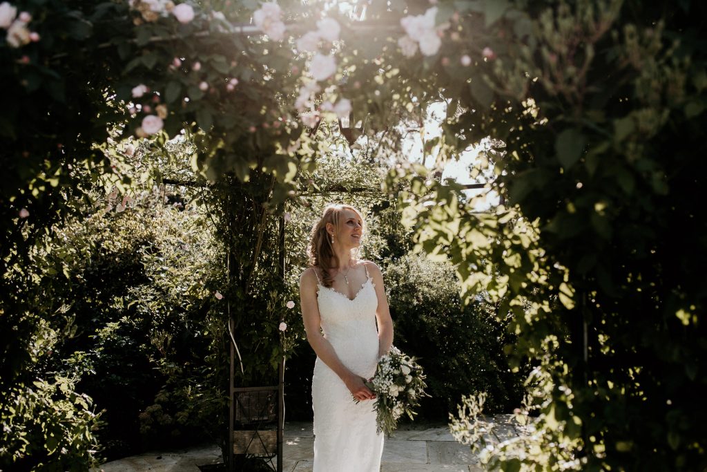 bride in luxurious white lace wedding dress with bouquet standing in the sunshine surrounded by trees and rich foliage