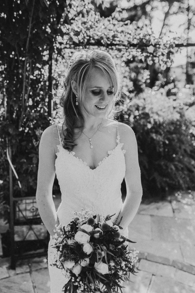 black and white close up of bride in luxurious white lace wedding dress with bouquet standing surrounded by trees and rich foliage