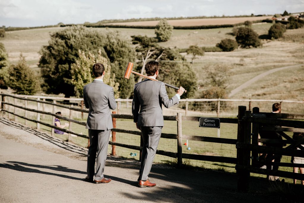 groom and groomsman in matching grey suits stand with croquet mallets and look over rolling english countryside