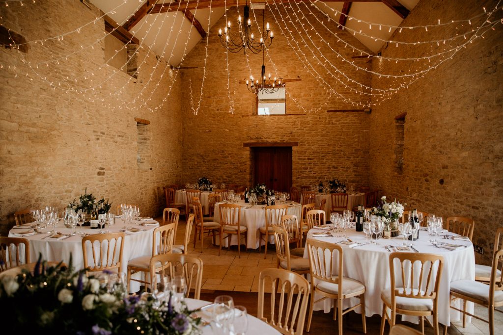 luxurious beamed barn conversion wedding venues laid out for reception with wooden chairs, fairy lights and tabled laid with white table cloths and floral centre pieces and glassware