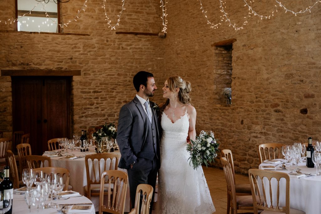 bride and groom smile at each other in luxurious beamed barn conversion wedding venues laid out for reception with wooden chairs, fairy lights and tabled laid with white table cloths and floral centre pieces and glassware