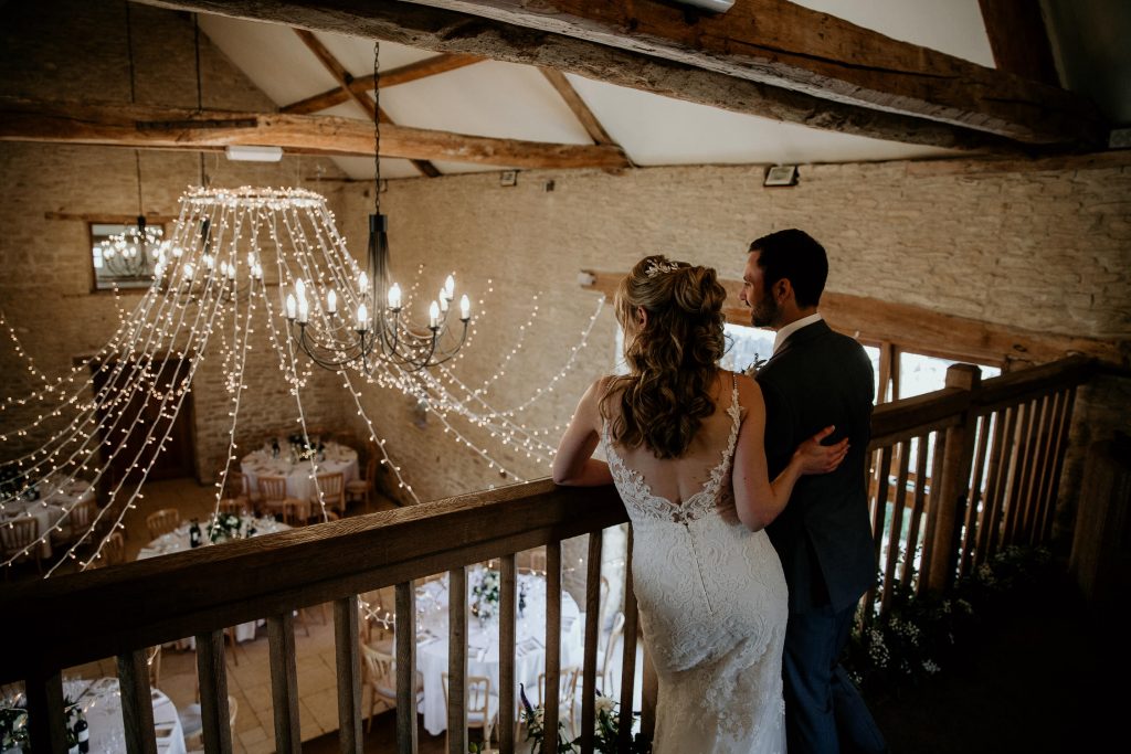bride and groom stand on balcony overlooking luxurious beamed barn conversion wedding venues laid out for reception with wooden chairs, fairy lights and tabled laid with white table cloths and floral centre pieces and glassware