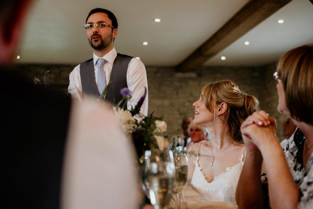 seated bride in intricate lace wedding dress looks up at groom in grey waistcoat and light blue tie making wedding speech