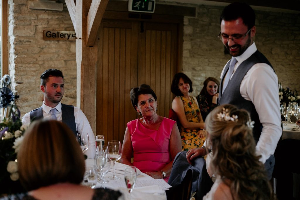 groom in grey suit with light blue tie looking down at bride whilst making wedding speech with smiling guests around them