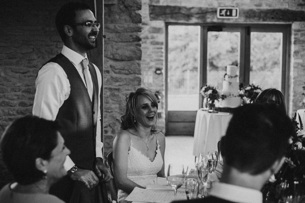 black and white photo showing groom in suit giving wedding speech and bride laughing seated beside him