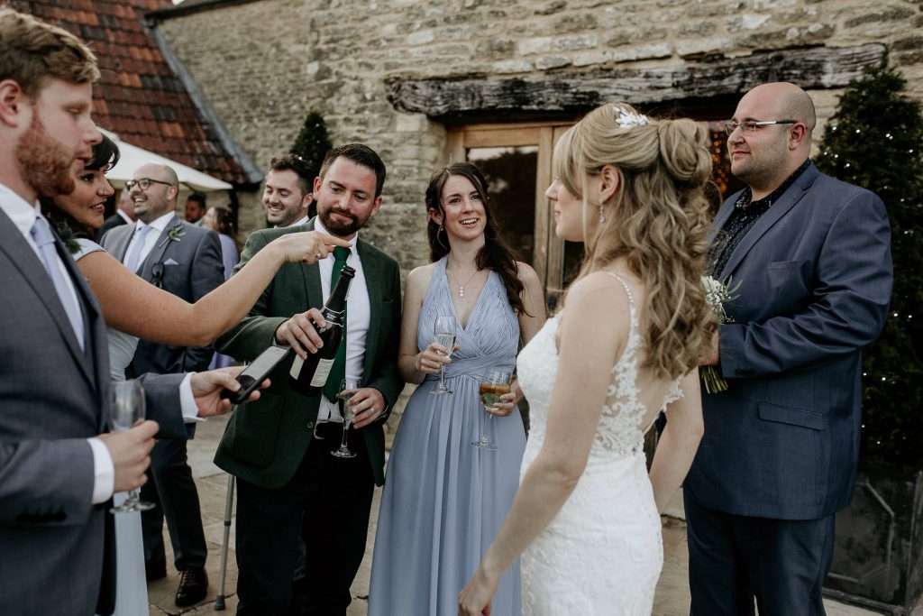 bride indelicate lace white wedding dress greets wedding guest celebrating with champagne outside of luxurious barn conversion wedding venue