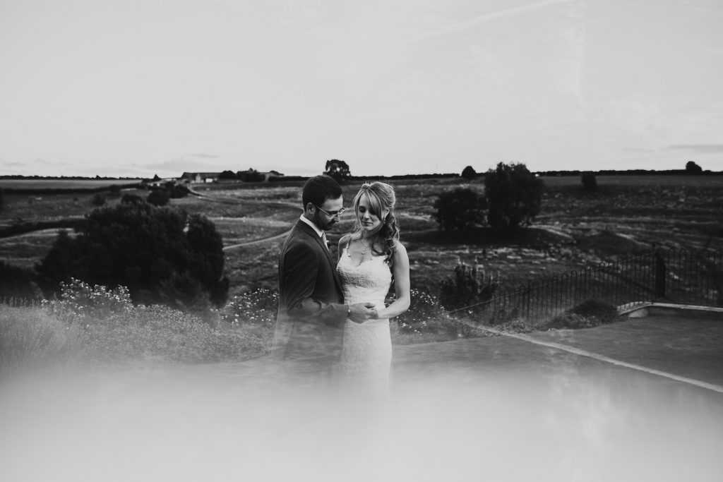 bride wearing intricate lace white wedding dress stands with groom in grey suit with beautiful rolling english countryside in the background black and white