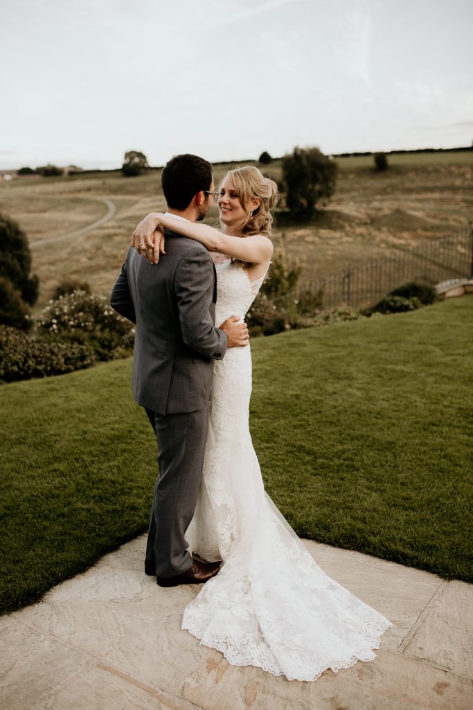 bride wearing indelicate lace white wedding dress hugs groom in luxurious grey suit with beautiful rolling english countryside in the background