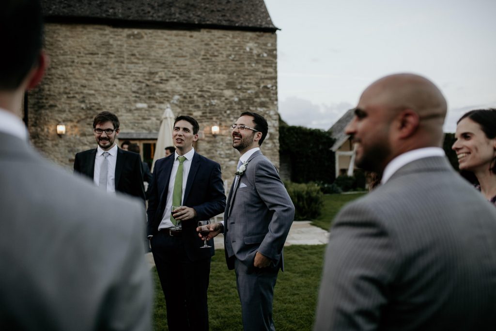 groom in grey suit with white rose buttonhole stands laughing with guests with luxurious barn conversion kingscote barn wedding venue in the background