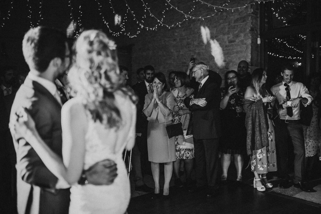 black and white sentimental photo of wedding guests smiling as they watch the bride and groom take their first dance