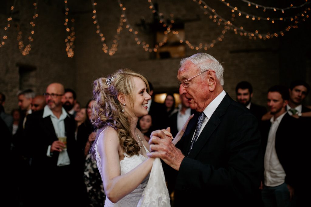 bride in white wedding dress dances with elderly wedding guest with other wedding guests in the back ground