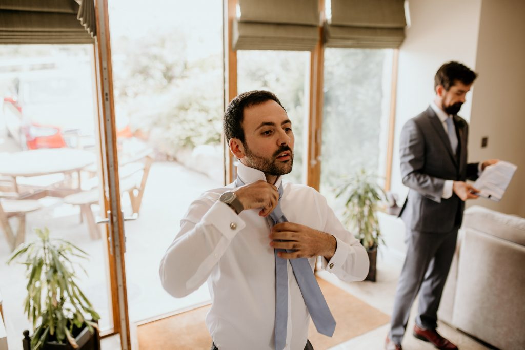groom stand in front of sunny glass doors doing up his luxurious light blue tie