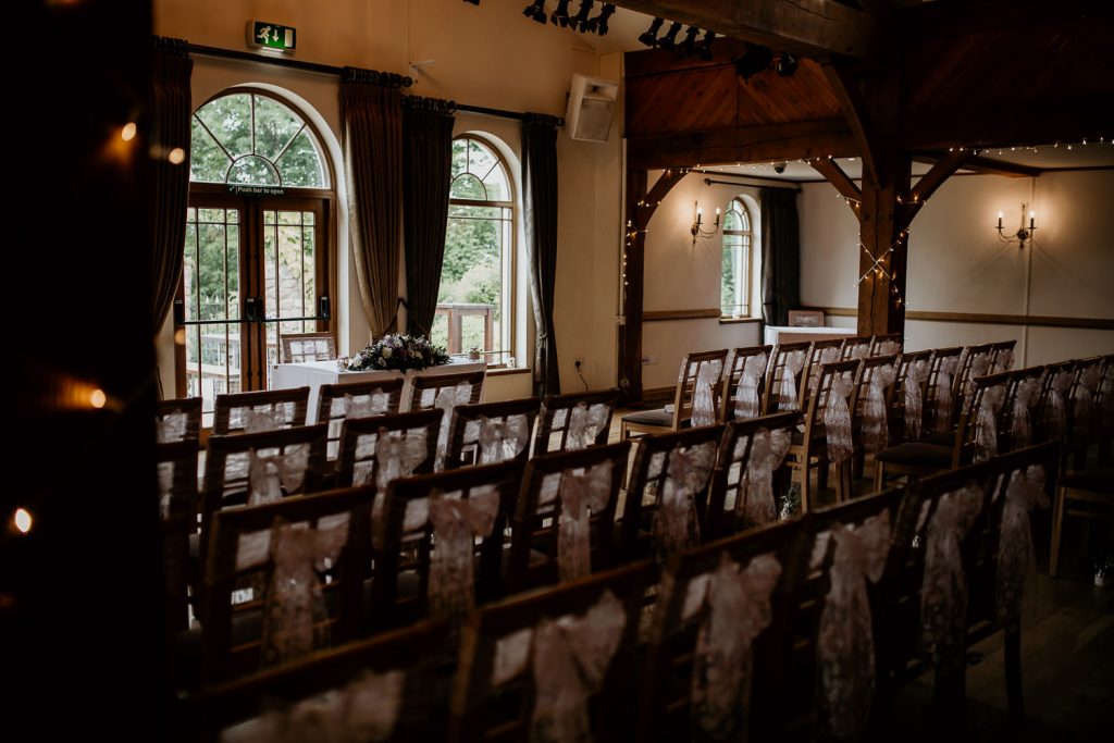 elegant wedding ceremony venue with wooden chairs with ribbons and fairy lights on the beams