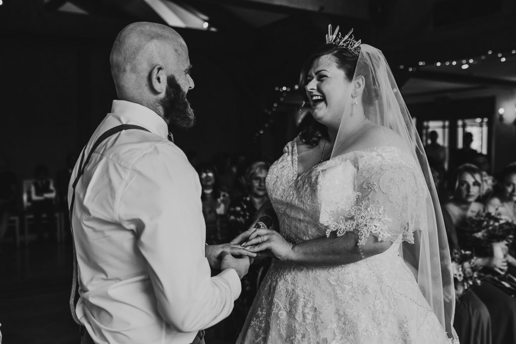 black and white photo of happy and smiling bride placing ring on grooms finger