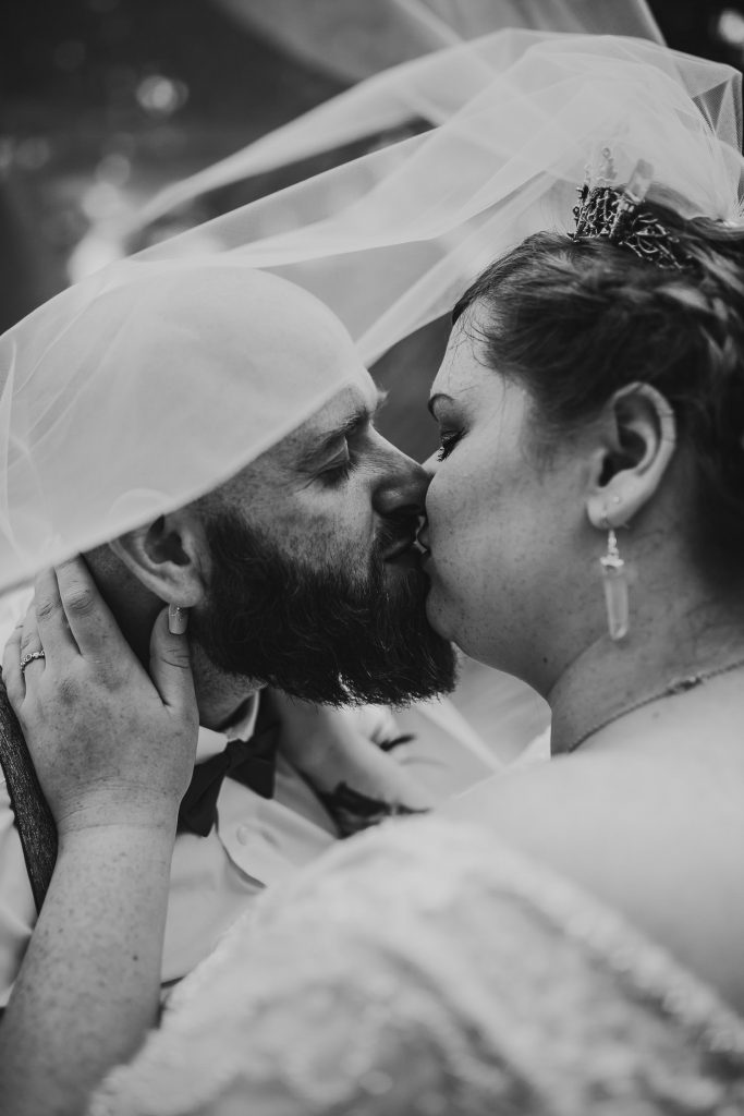 black and white photo of quirky alternative wedding bride and groom kissing under flowing veil