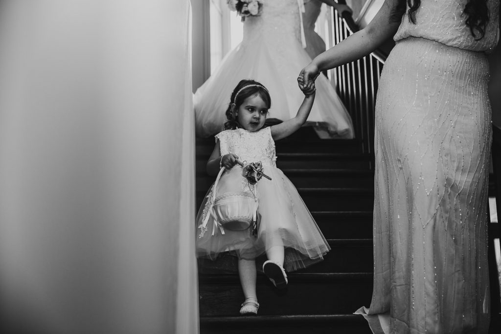 black and white photo of young flower girl walking down stairs with bridesmaid in front of the bride