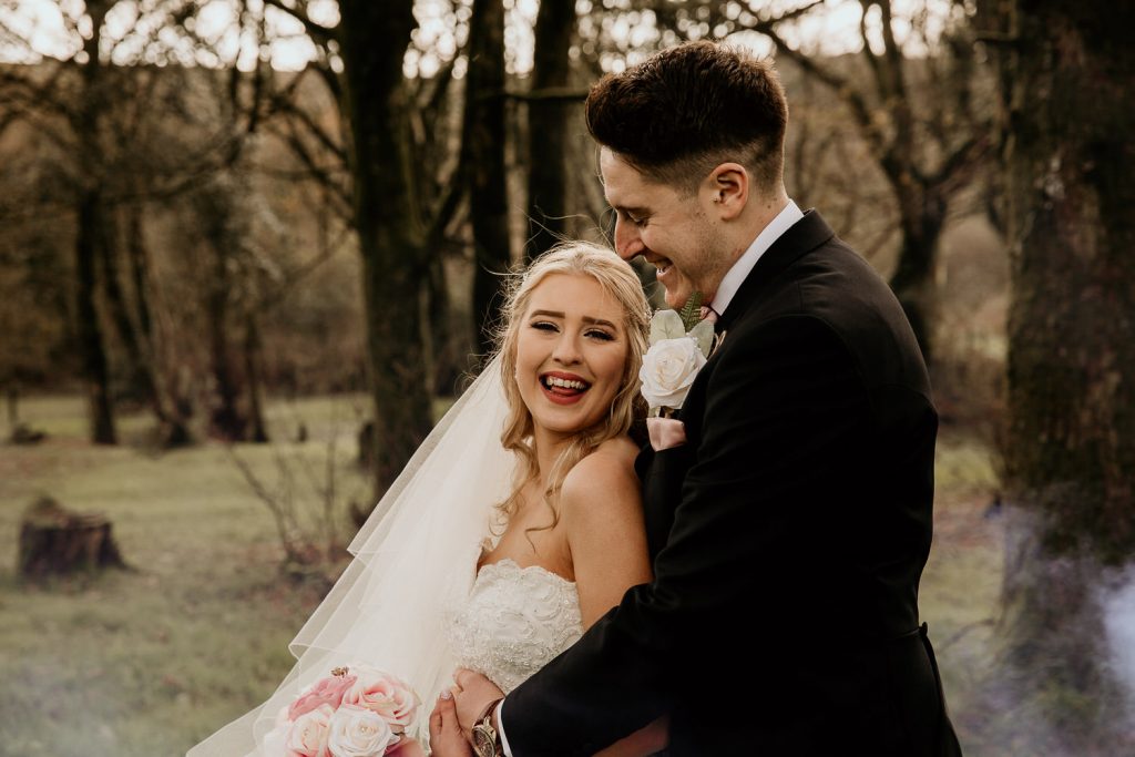 laughing bride and groom hug outside of venue in wooded area