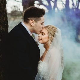 groom kisses brides forehead in smokey wooded area