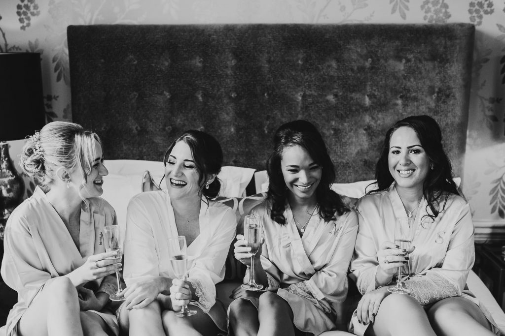 bride and bridesmaids in matching luxurious white dressing gowns laugh and drink champagne