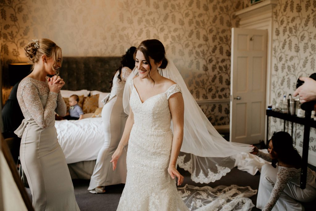 smiling bride and bride and bridesmaid get ready for the wedding in grand looking hotel room with luxurious cream and gold wallpaper