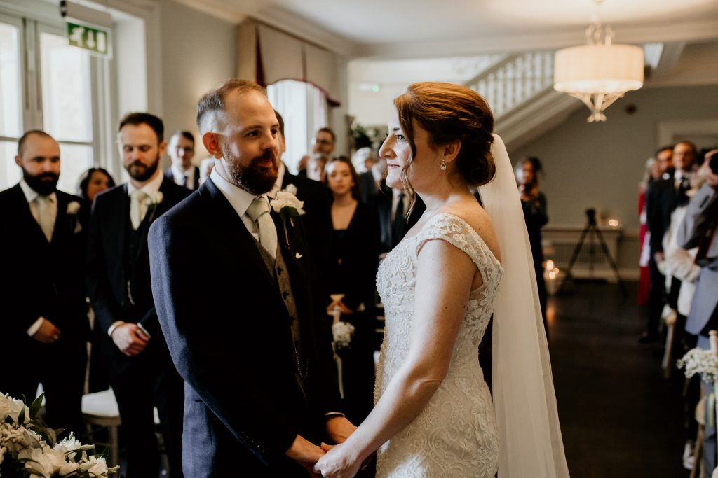 bride in embroidered wedding dress and groom in chic black suit with grey waistcoat, green tie and white rose buttonhole stand facing each other hand in hand taking their vows