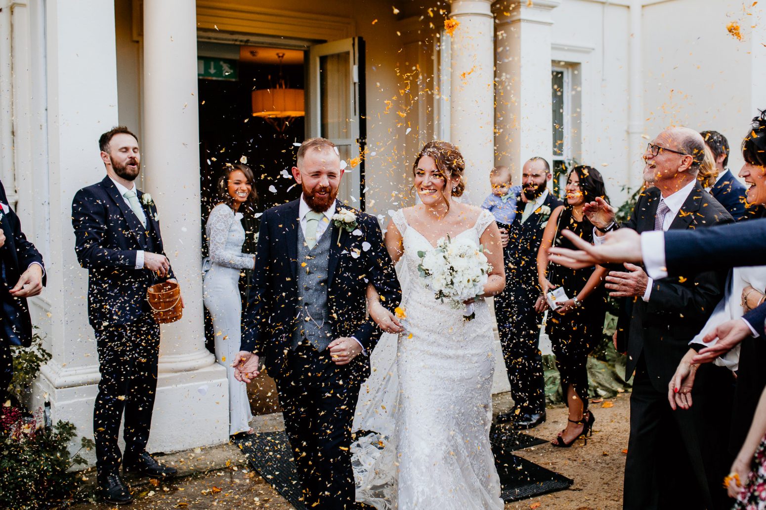 bride in embroidered wedding dress and groom in luxurious suit with green tie and white rose buttonhole walk arm in arm under falling confetti surrounded by smiling wedding guests