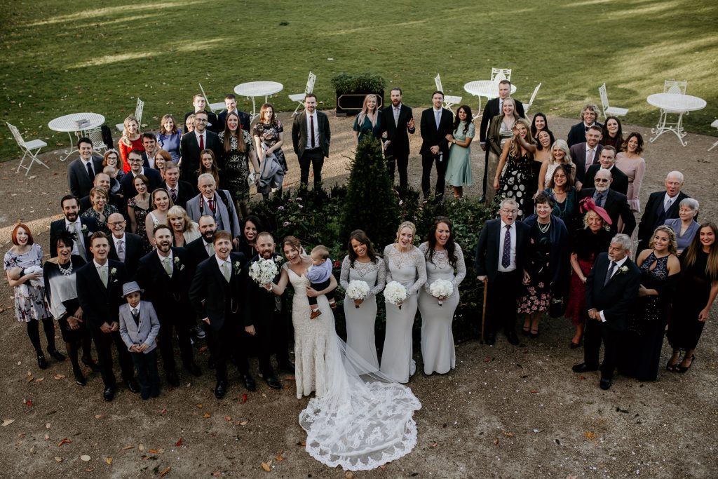 bride in embroidered wedding dress and groom in luxurious suit with green tie and white rose buttonhole stand with wedding party