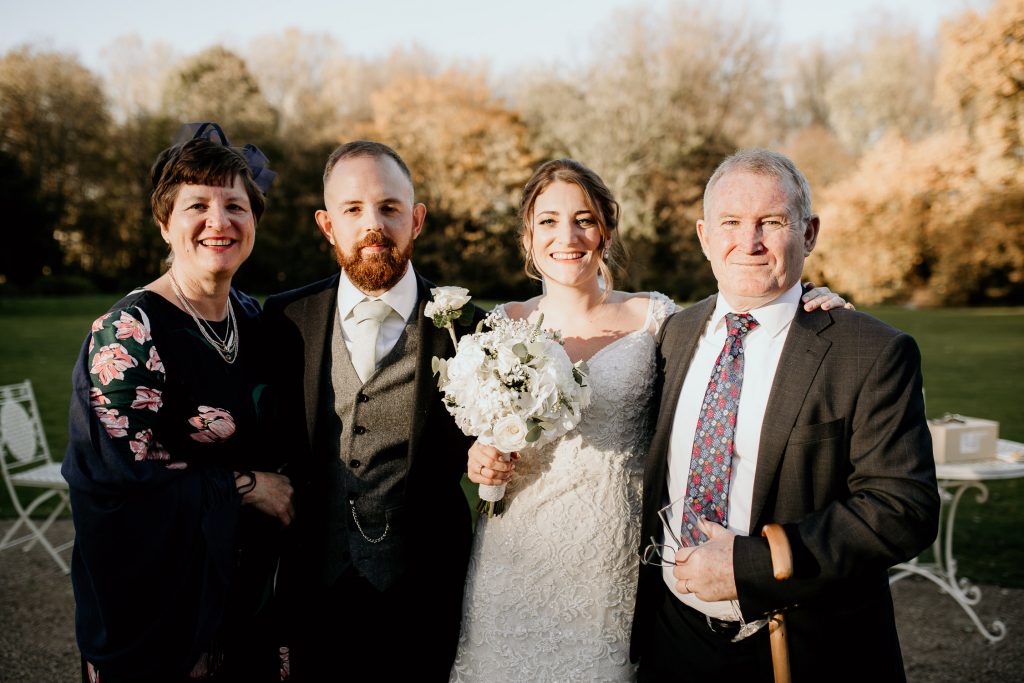 bride and groom groom stands outside of wedding venue in the sunshine with wedding party