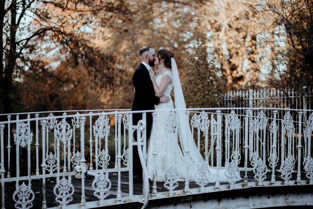 bride and groom kiss on beautiful white bridge with trees and delicate metal fence in the background