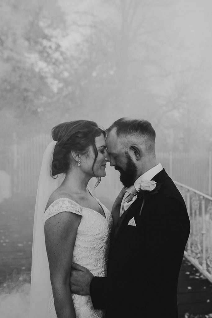 black and white smokey photo of bride and groom embracing on beautiful white bridge with trees and delicate metal fence in the background