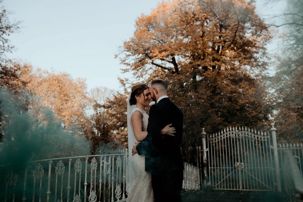 black and white smokey photo of bride and groom embracing on beautiful white bridge with autumn trees and ornate white gate and fence in the background