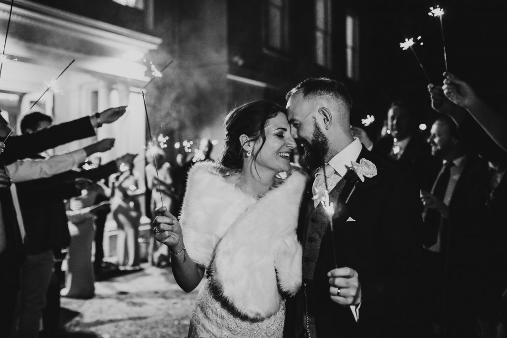 black and white photo of bride and groom laughing and hugging outside of wedding venue in the night with wedding guests surrounding them with sparklers