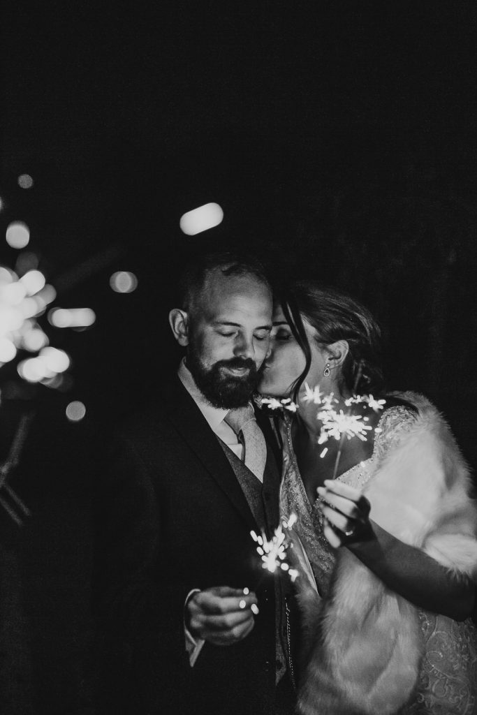 black and white photo bride kisses groom on the cheek whilst they both old sparklers outside of wedding venue in the night time