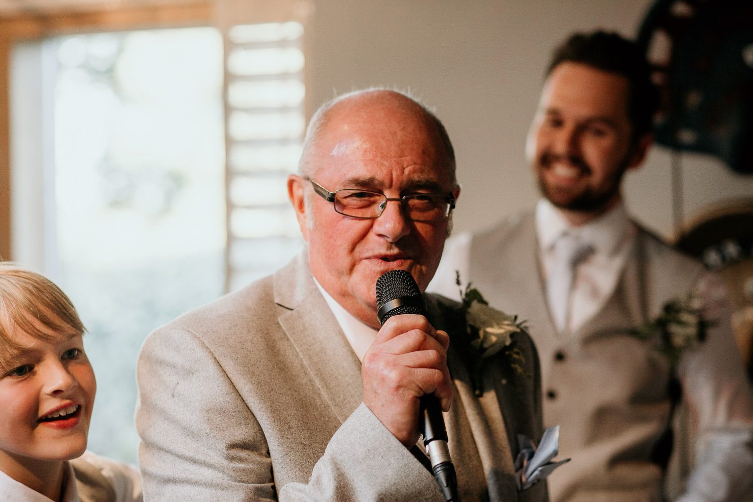 man in grey suit gives speech whilst groomsman and young boy smile