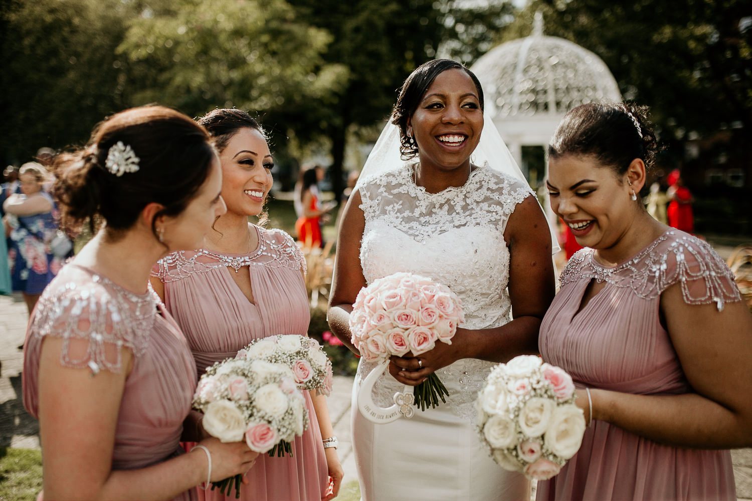 happy photo of bride in white dress and bridesmaids in pink all with white and pink bouquets laughing together outside in the sun