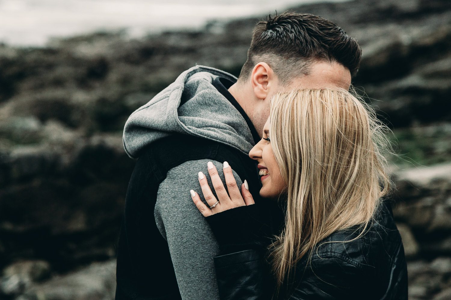 Laughing bride and groom cuddle windy seaside engagement shoot