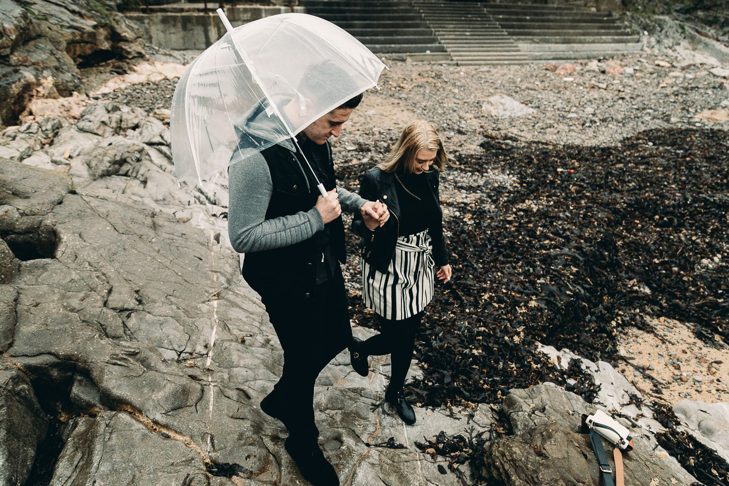 Bride and groom walk hand in hand under umbrella during seaside engagement soot